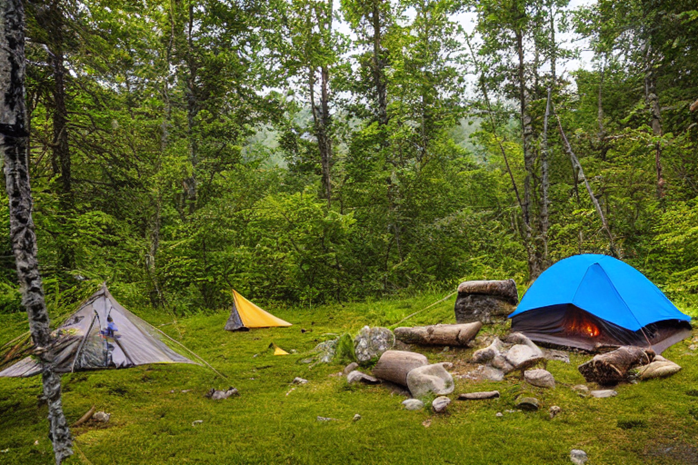 A tan tent rests in a peaceful valley as the sunrise warms the clear sky. A stone-ringed campfire, cozy hammock, and small stream make this campsite feel calm and inviting.
