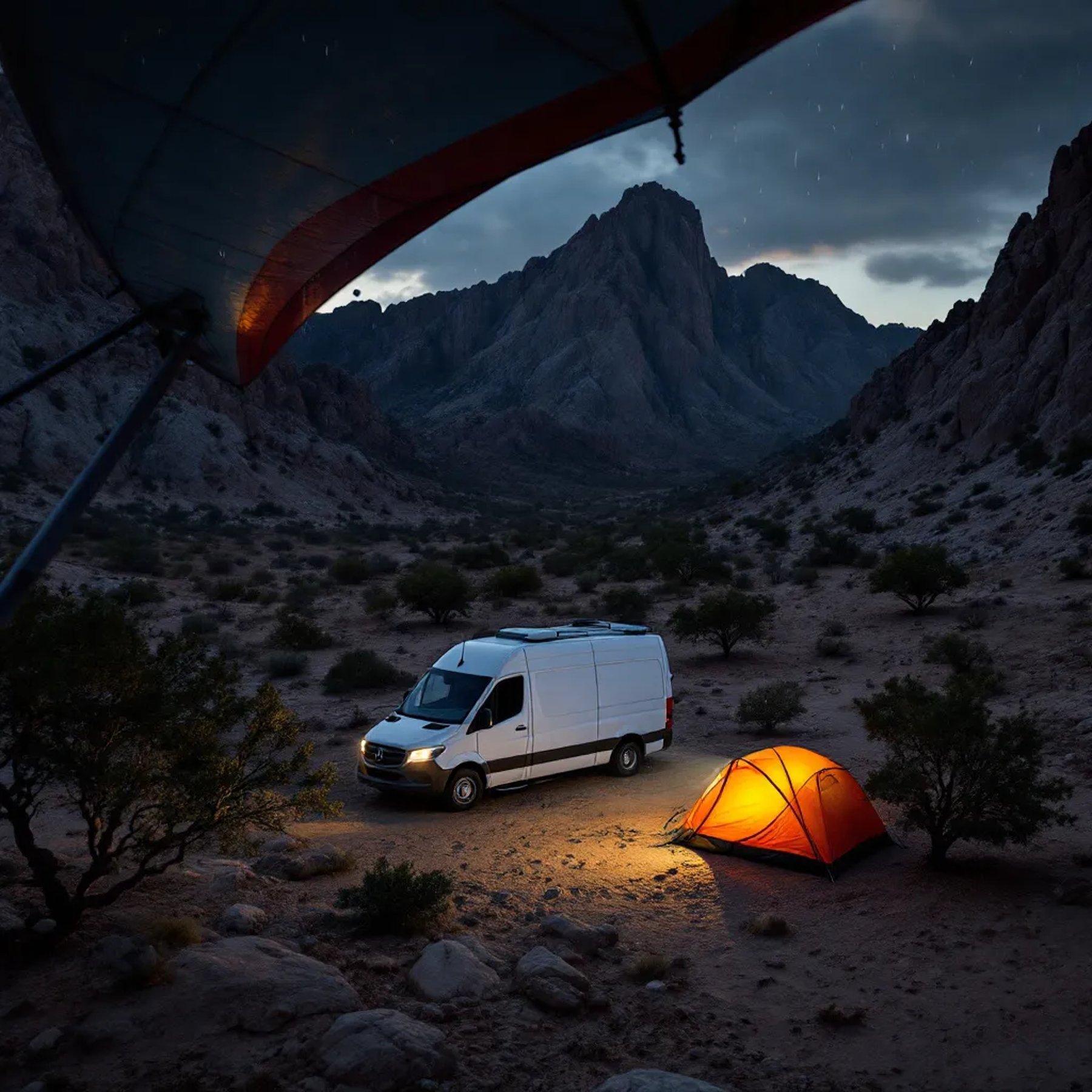 A white camper van rests among big rocks and small trees in Big Bend at dusk. A nearby tent glows softly, while animal tracks mark the dusty ground. Dark clouds bring a wild, dramatic feeling to the quiet campsite.