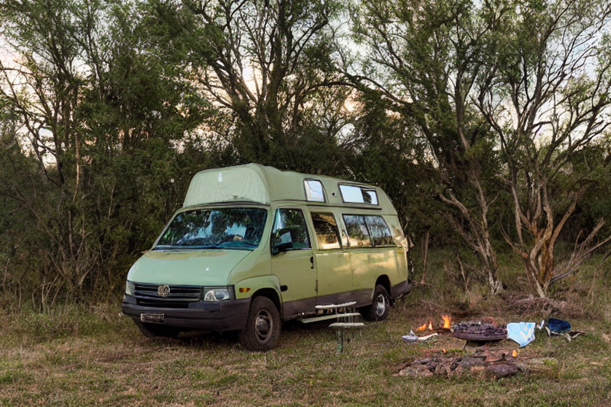 A green camper van rests on a rocky campsite as the sun sets over Big Bend’s wild desert hills. A warm campfire glows nearby, with clothes hanging on a line and a deer standing in the distance beneath the clear sky.