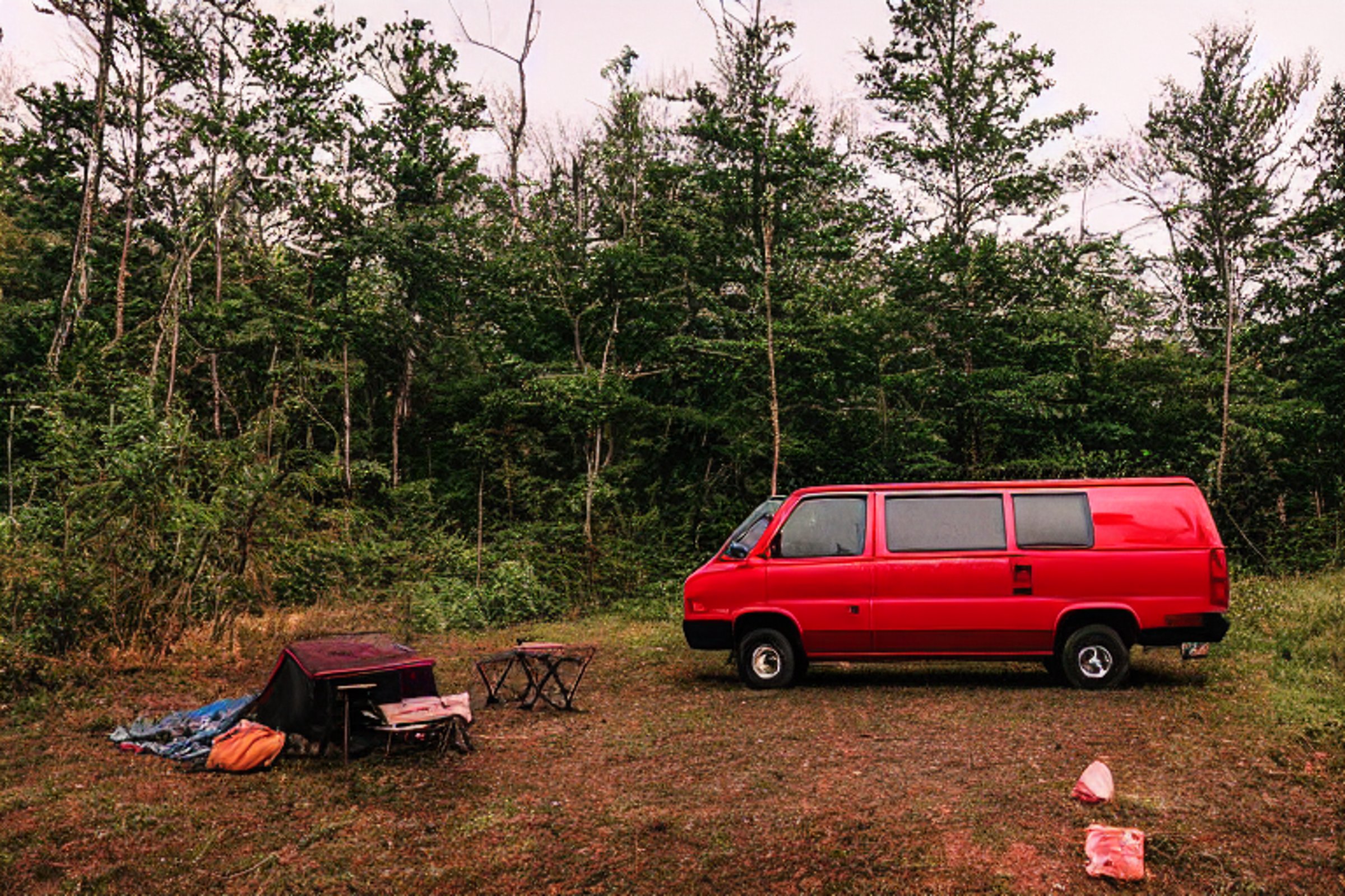 A red camper van rests at a paved campsite in the misty Smoky Mountains. Camping chairs and a guitar sit nearby as a hiking path winds into the trees. In the distance, a deer wanders through the soft golden rain.