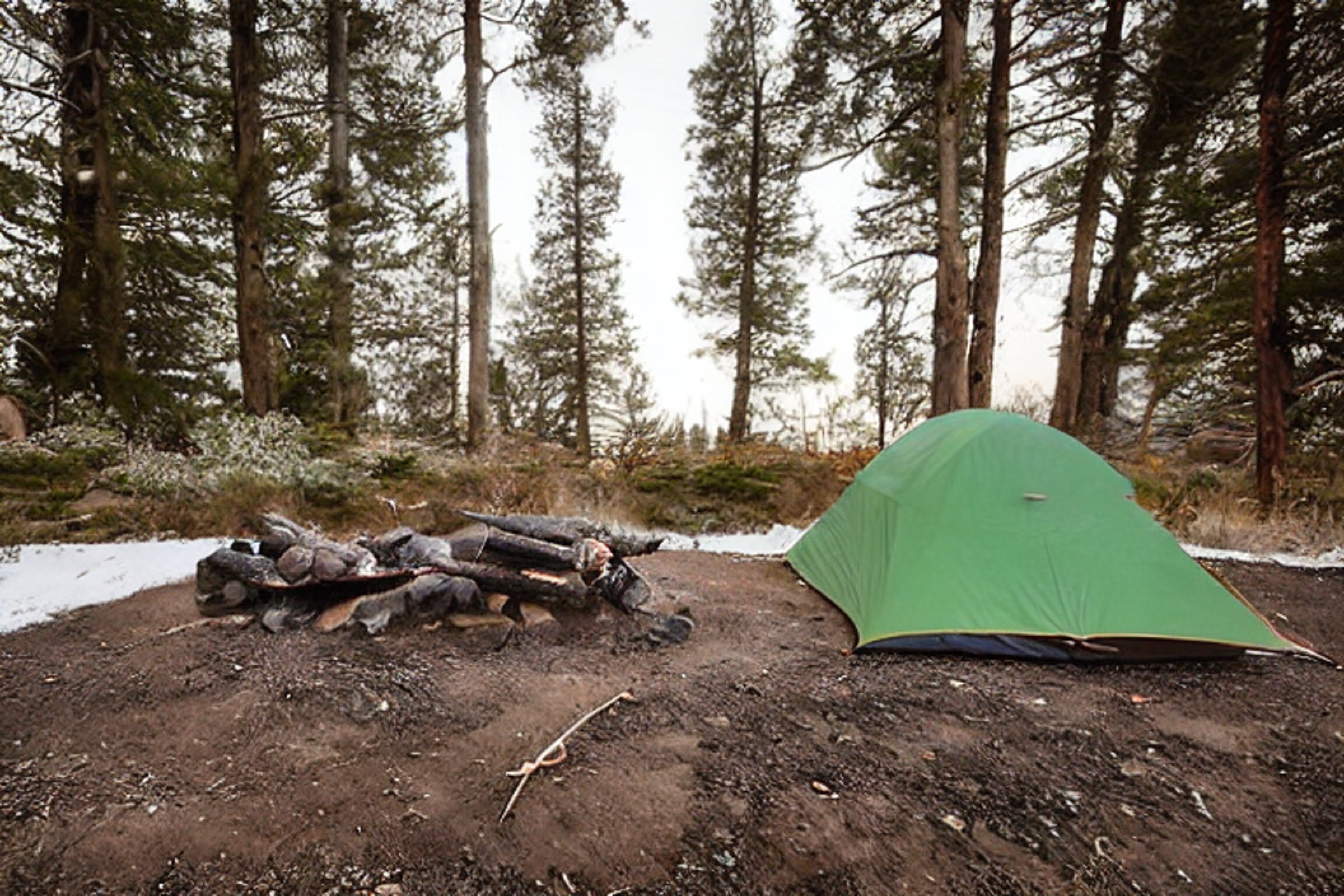 A green and tan tent rests on a rocky campsite along the California coast. Bushes and boulders surround the area as snow falls softly at dusk. A warm campfire glows beside the logs in the center.