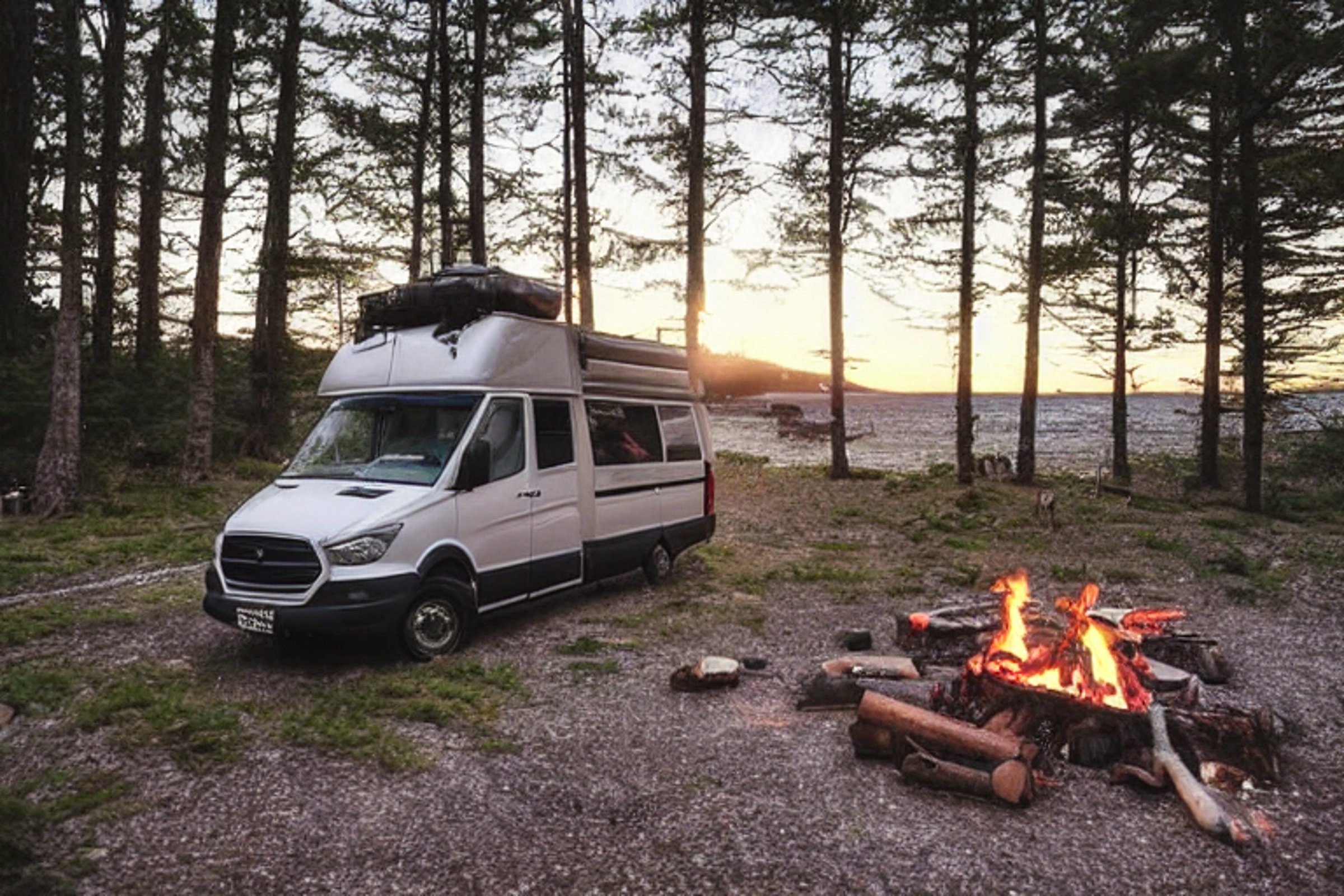 A cozy van campsite rests on a quiet beach beside the ocean. A warm campfire glows among the stones as the sun sets, with large rocks and gravel patches adding to the peaceful scene.
