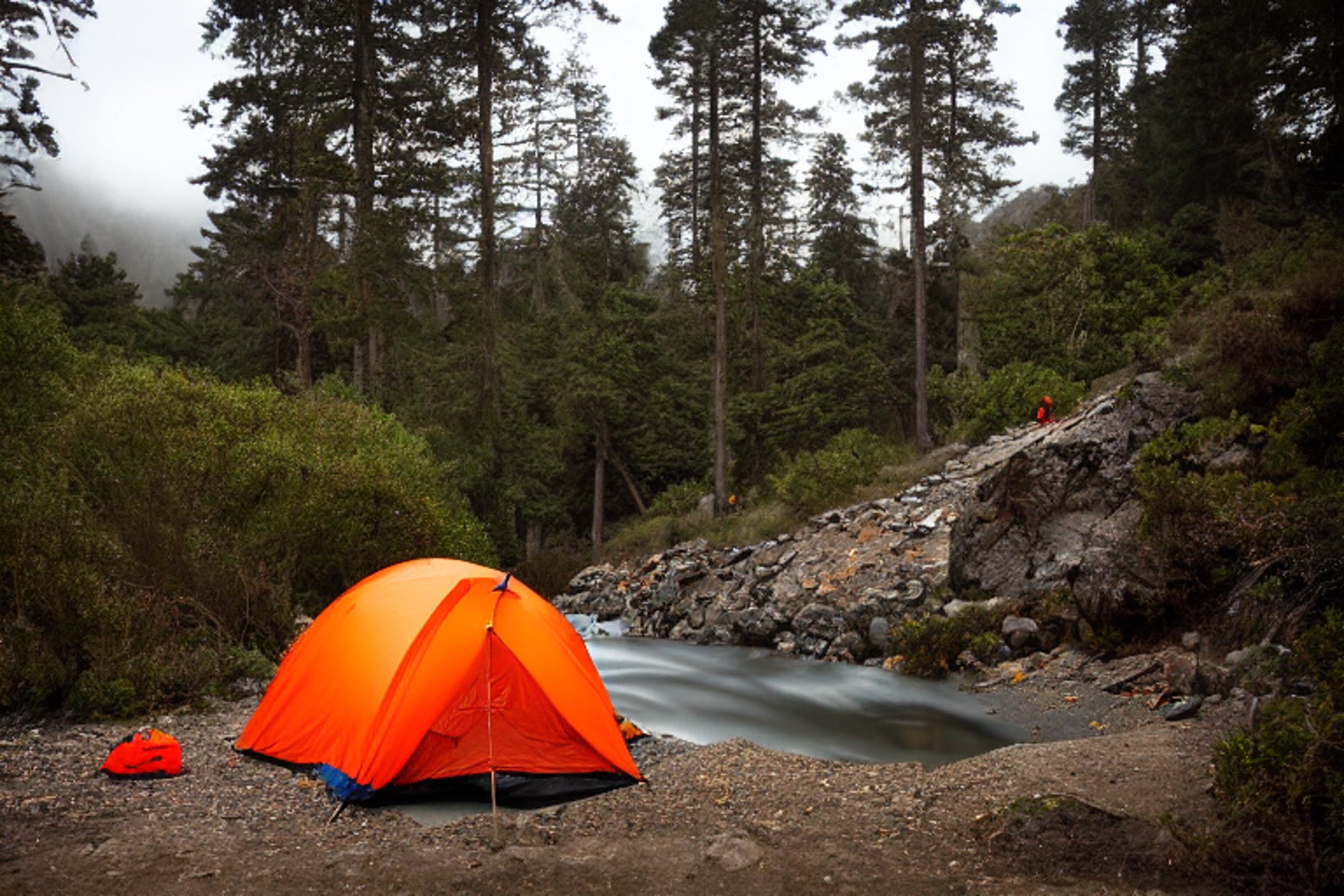 An orange tent rests on a rainy campsite in Big Sur at night. A small stream winds through the gravel and dirt, while a box of camping gear sits nearby. In the distance, a camper’s flashlight glows softly.