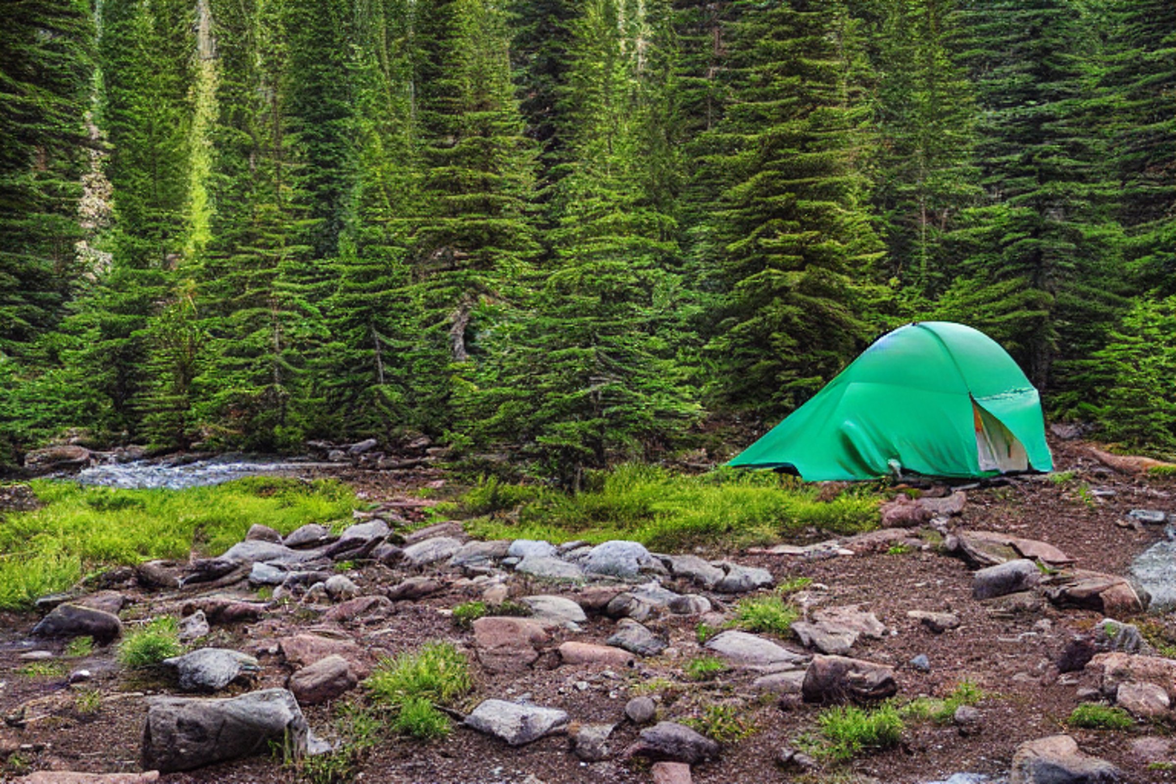 A green tent rests on a paved campsite in Glacier National Park. A clear stream winds past boulders and tall trees, while storm clouds glow in the sunset.