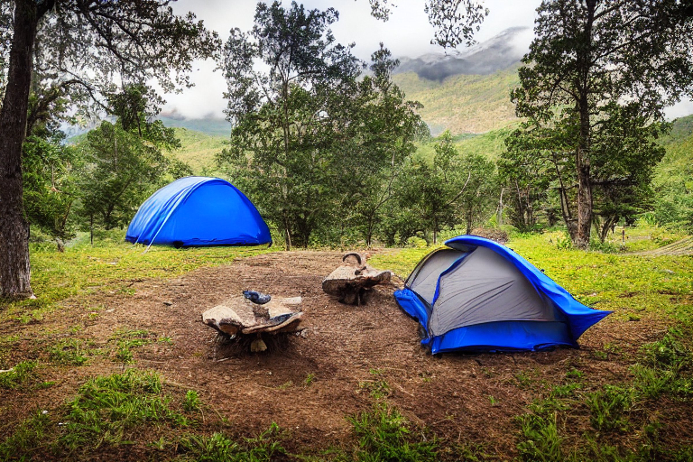 A blue tent rests beside a warm campfire on a dirt clearing near a quiet river. A hammock hangs between the trees, while a mountain rises in the distance under a stormy sunset sky. Fallen leaves cover the ground around the peaceful campsite.