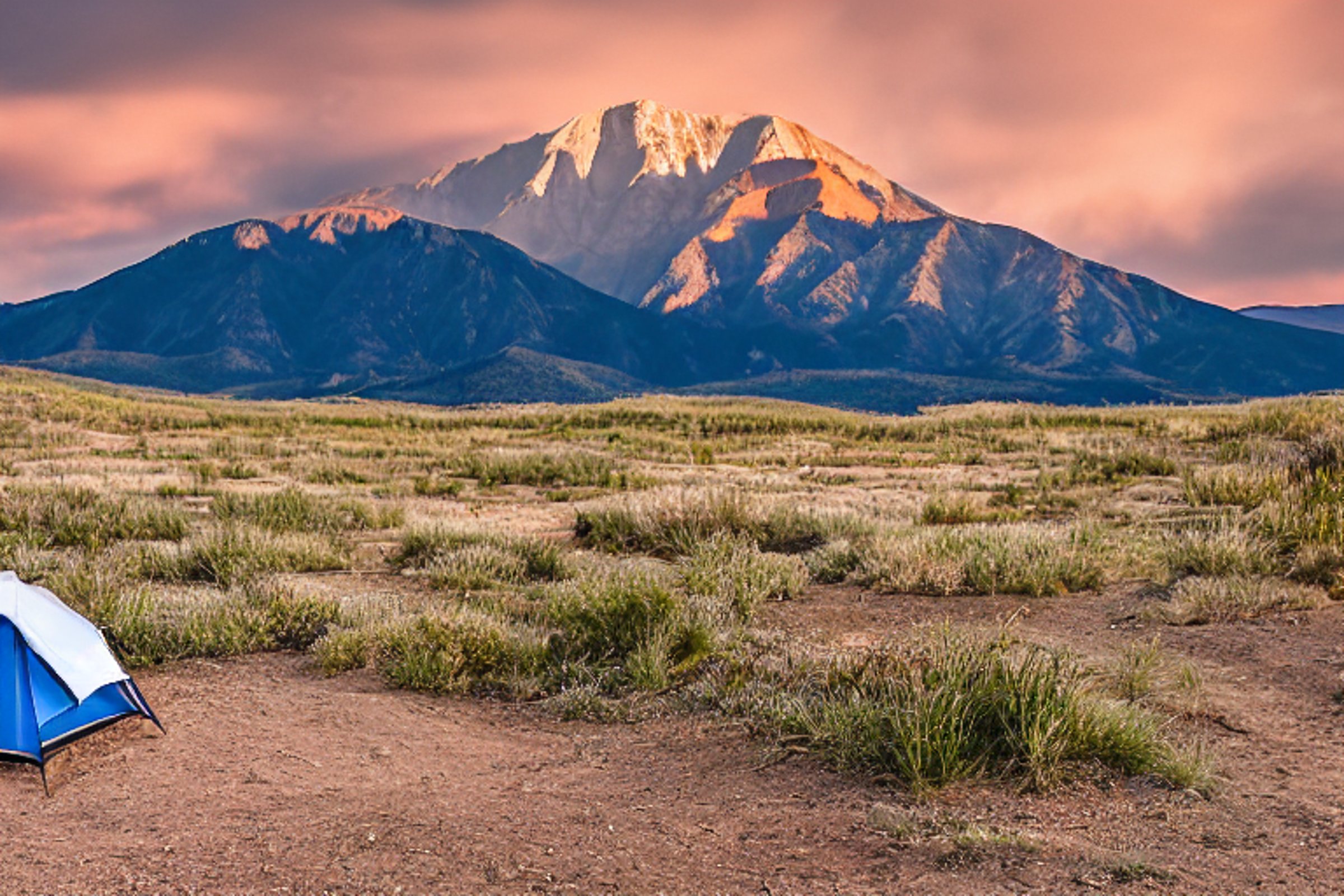 A bright blue tent rests on a rocky dirt patch beneath a rainy sunset sky. A warm campfire glows nearby, with small flowers and mountains rising in the distance.