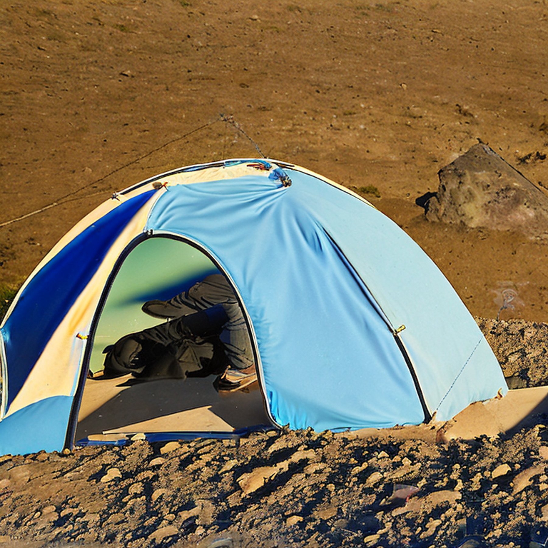 A gray and blue tent rests on a rough dirt campsite beneath a clear night sky. The colorful northern lights glow above the rocky ground, while a backpack and large stone slabs sit nearby.