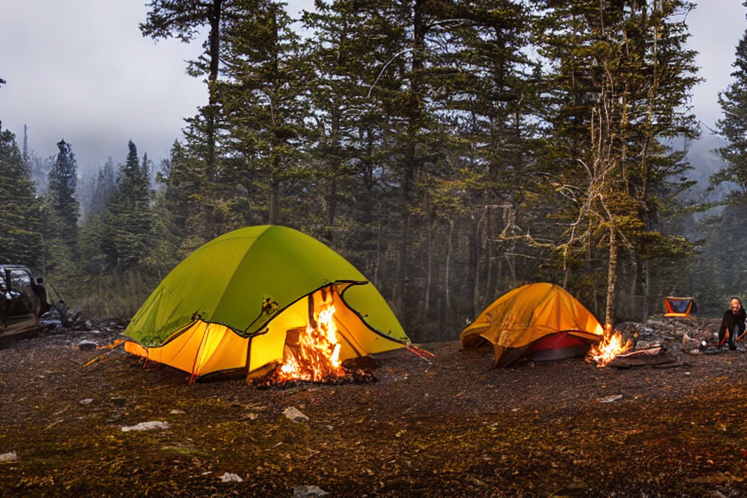 A forest-green tent rests on a paved campsite in the Smoky Mountains. Snowy fall trees glow beneath the colorful northern lights, while a warm campfire crackles inside a ring of stones.