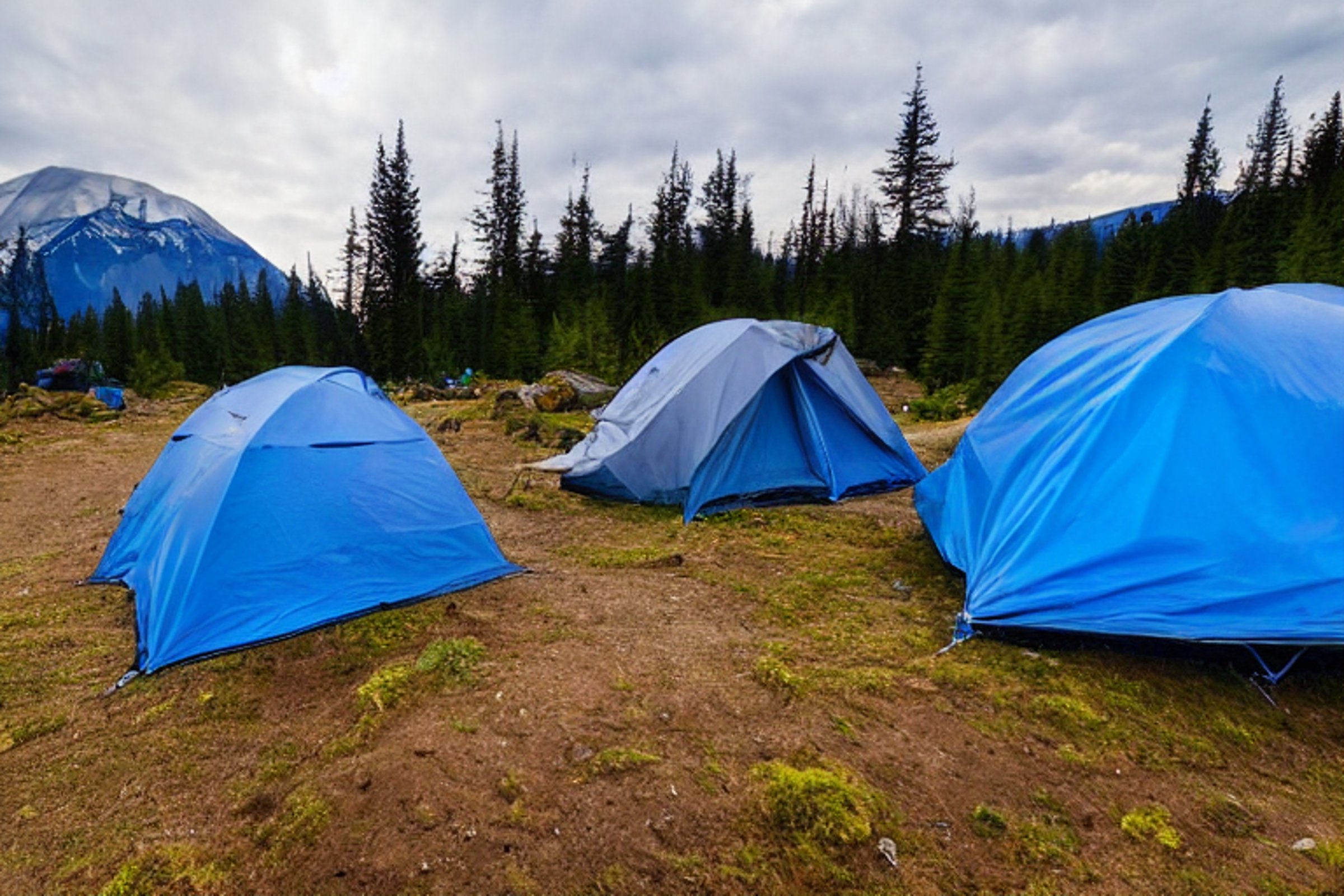 A blue tent rests on a quiet dirt campsite beneath a star-filled, cloudy sky. A mountain rises in the distance, while a clear stream winds past the camp chairs and hiking path.