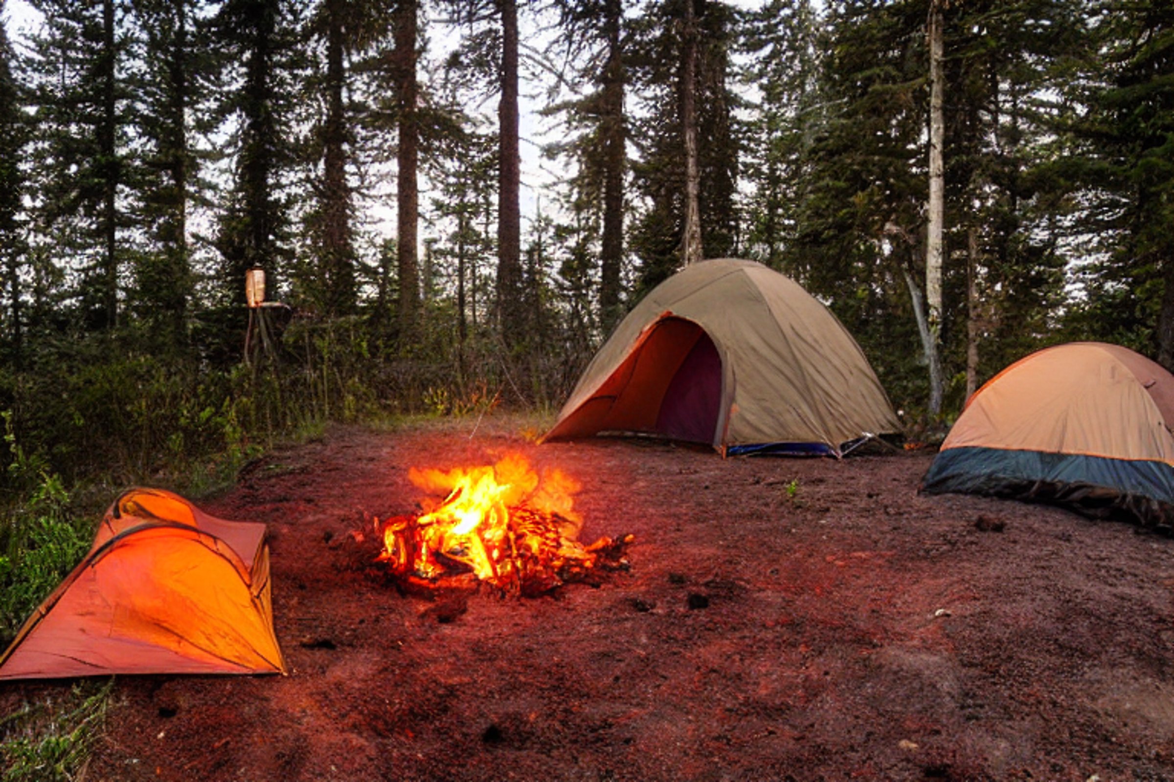 A cozy brown tent rests beside a paved campsite on the rainy California coast. A stone-ringed campfire, glowing lantern, and clothesline add a warm, homey feel. Soft sunrise light shines through the cloudy sky.