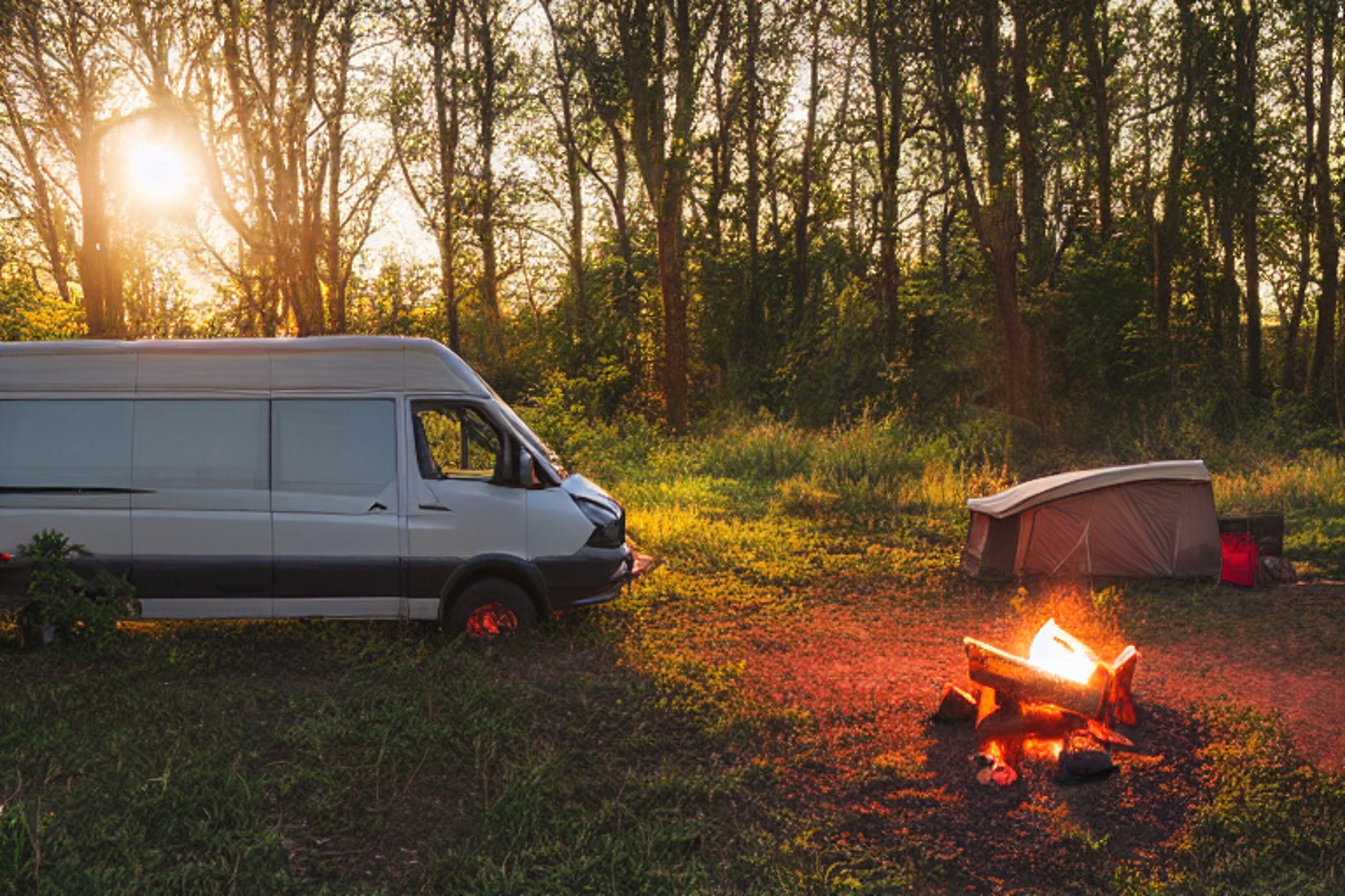 A cozy camper van rests beside a quiet river as the sun sets. A bright campfire glows on the dirt, with logs, fallen leaves, and a lantern nearby. Berry-covered bushes add a splash of color to this peaceful campsite.