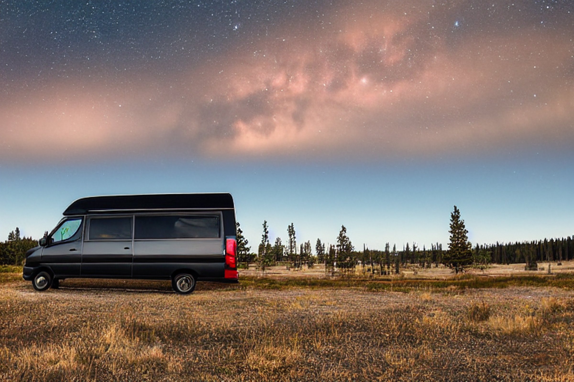 A black camper van rests at a paved campsite in Yellowstone as the sun rises. Camping gear sits nearby, while a dirt patch and open landscape stretch toward the horizon. A pale moon hangs in the clear morning sky.