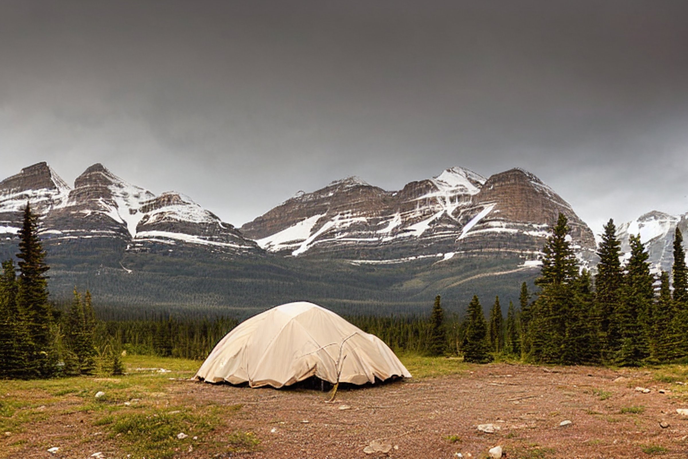 A cozy brown tent sits on a paved campsite in Glacier National Park. Tall bushes surround a dirt path and open patch of ground, with rolling hills beyond. Soft sunset light shines through the snowy air, creating a peaceful mountain mood.
