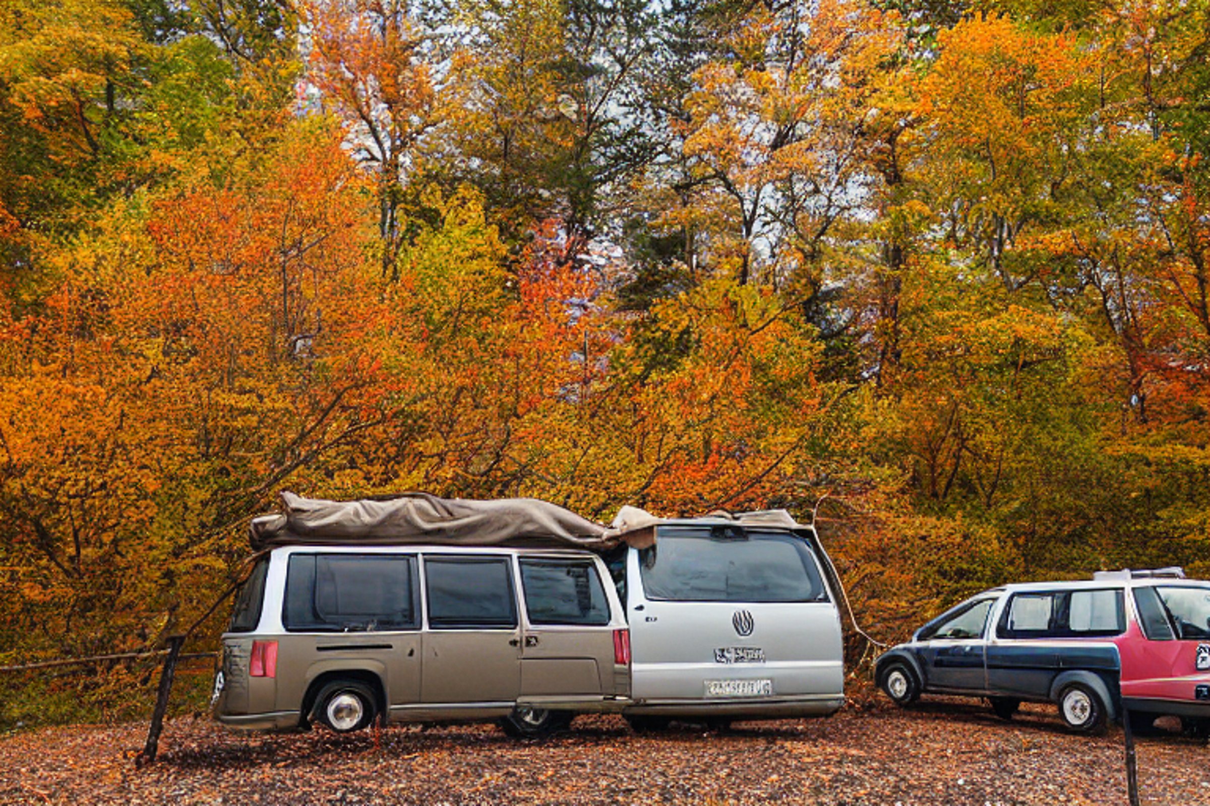 A cozy campervan rests beside a glowing campfire under stormy night skies. Fall-colored trees surround the dirt campsite, with a hiking path, picnic table, and clothesline nearby.
