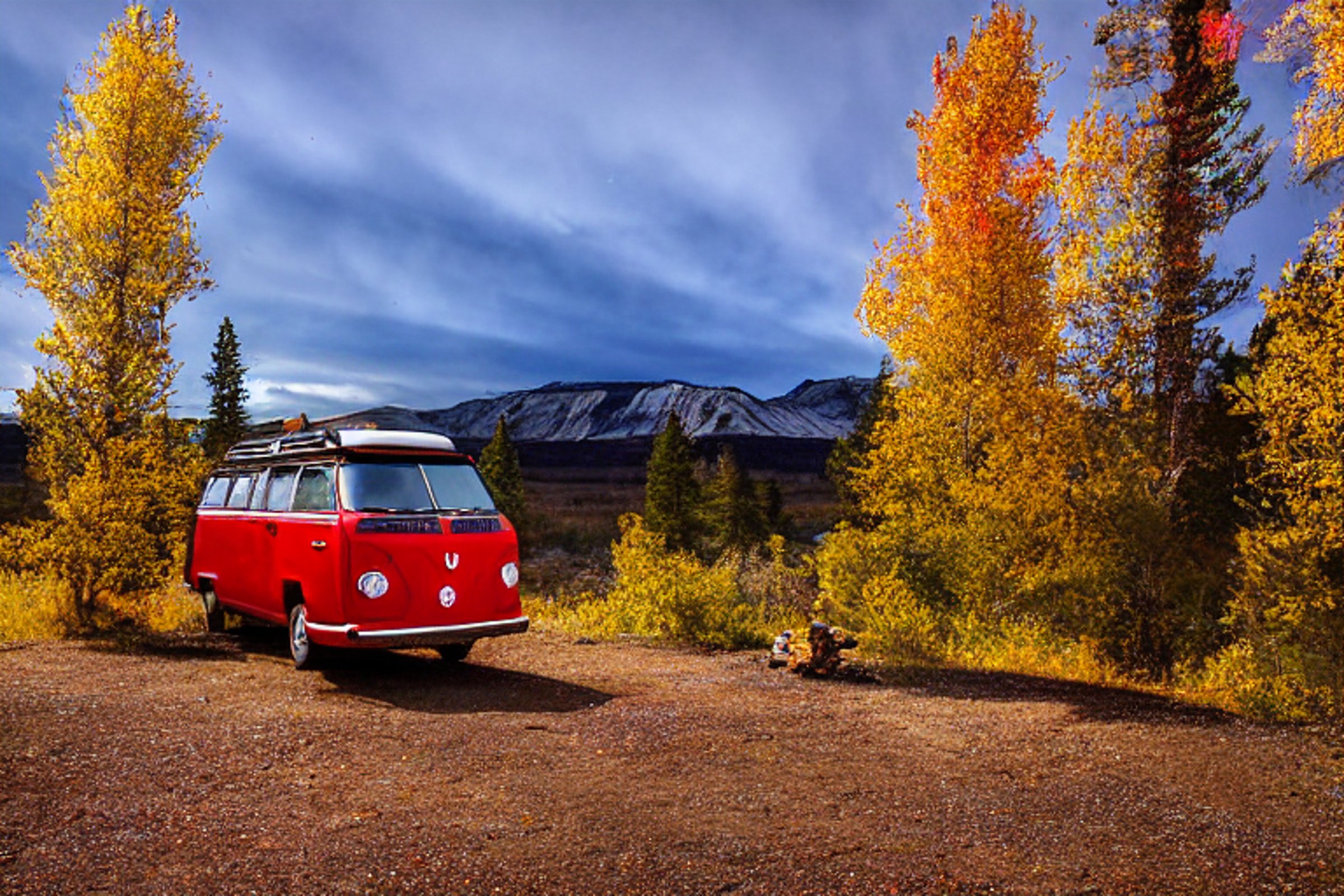 A red VW campervan rests at a snowy campsite beneath glowing green auroras. A warm campfire flickers beside the dirt clearing, with bright fall trees and tall mountains all around.