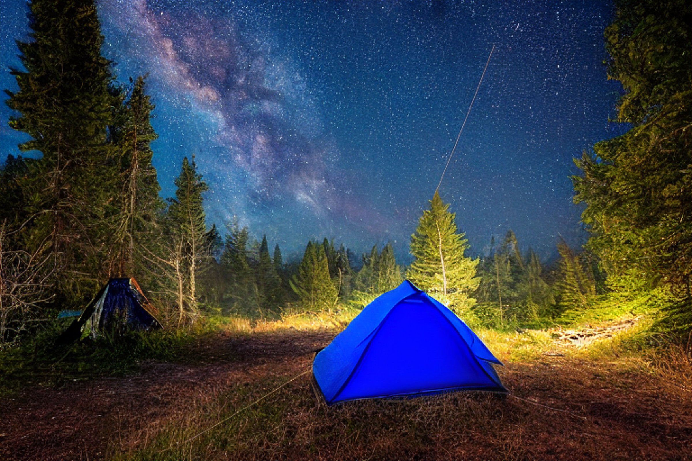 A bright blue tent glows beside a crackling campfire under a sky full of stars. Fog drifts over the rolling Washington hills, while clothes hang from a nearby line. A dirt path winds through the peaceful campsite.