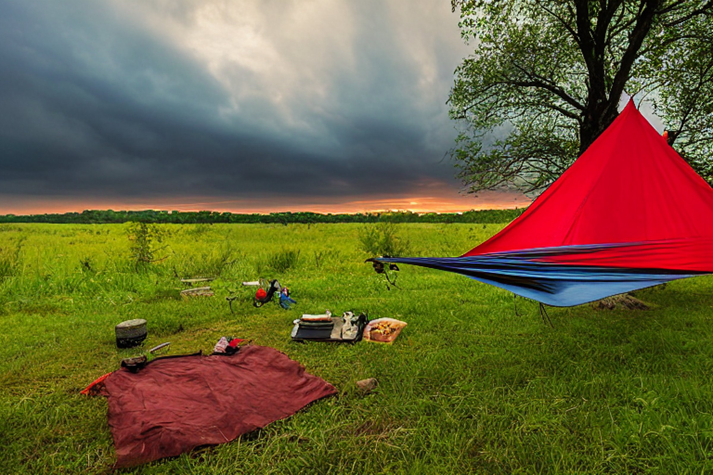 A red tent rests on a quiet dirt campsite as the sun sets over the rolling hills of Upstate New York. A hammock hangs between the trees, with camping gear nearby and a wooden guitar on the ground. Clouds soften the sky, creating a peaceful evening mood.